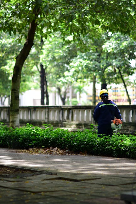 A gardener wearing a wide-brimmed hat, glasses, and a blue uniform with a badge on the sleeve is operating a handheld petrol-powered grass trimmer to trim the edges of a well-maintained lawn in a land